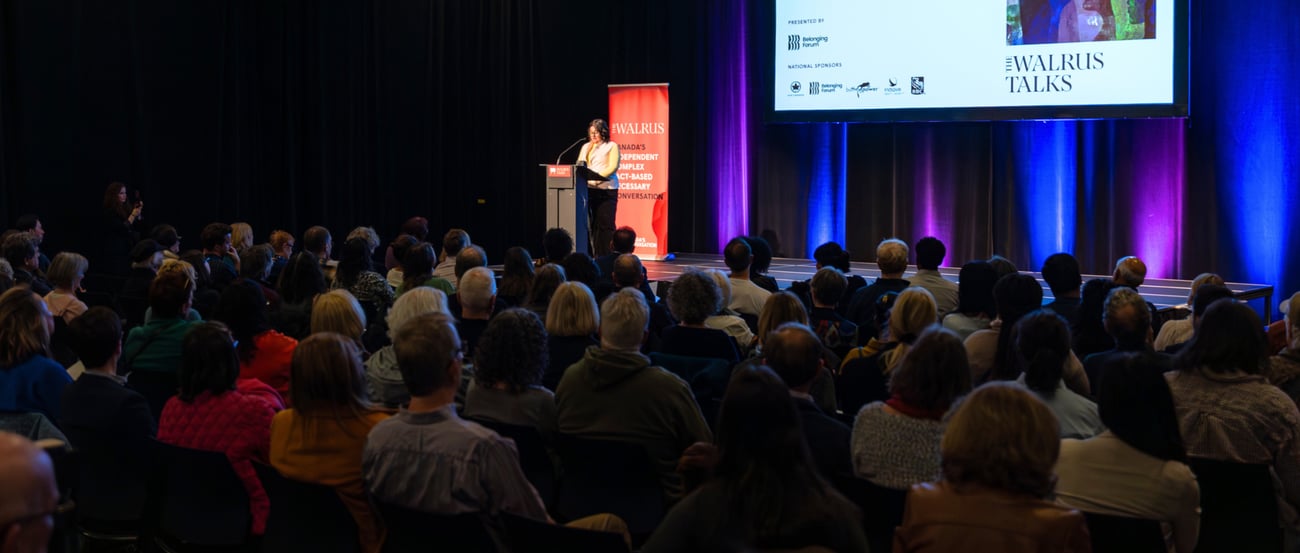 A speaker stands on stage at a podium, with a red The Walrus Talks banner behind her. In the foreground, the audience looks towards the stage, listening to her talk. 