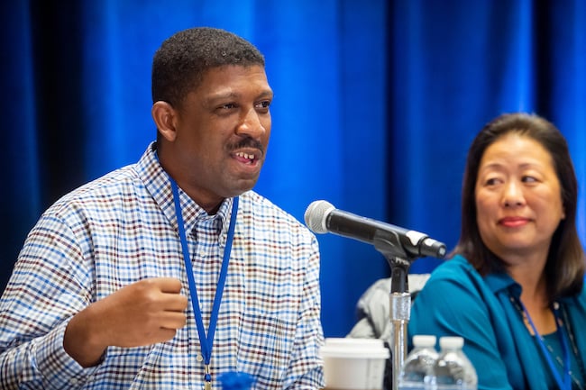 The image shows a panel discussion or conference setting. A man in a checkered shirt is speaking into a microphone, gesturing with his hand as he makes a point. He’s wearing a lanyard, suggesting he is a speaker or participant. Next to him, a woman in a teal top is seated, looking toward him attentively. On the table in front of them are water bottles and a takeaway coffee cup, and a blue curtain forms the background, typical of a formal event or conference stage.