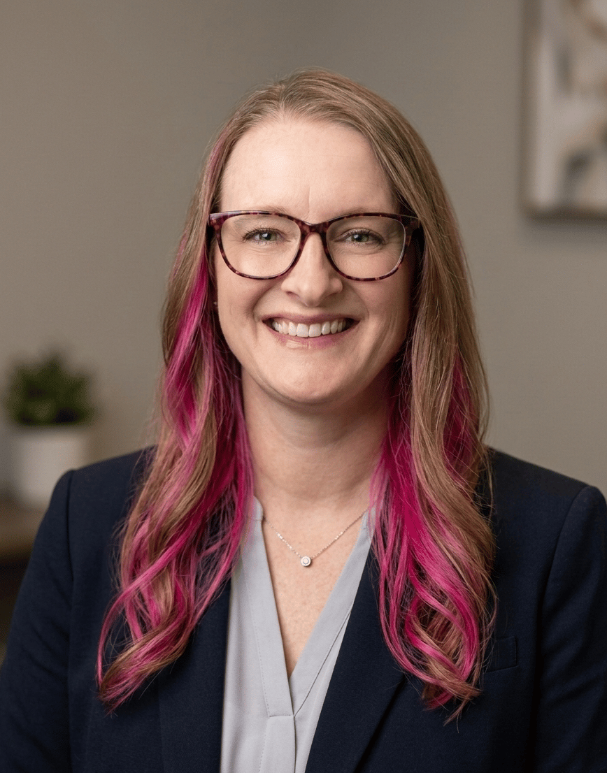 Julie Eshleman, a person with long wavy hair that transitions from light brown to bright pink at the ends is shown wearing a dark blazer over a light-colored top. The individual is positioned indoors against a softly focused background featuring neutral walls and a small plant on a table.