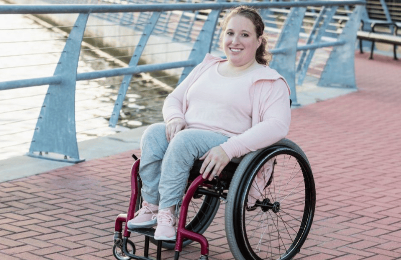 A young woman with spina bifida sits in her wheelchair and smiles. She is on a waterfront esplanade. 