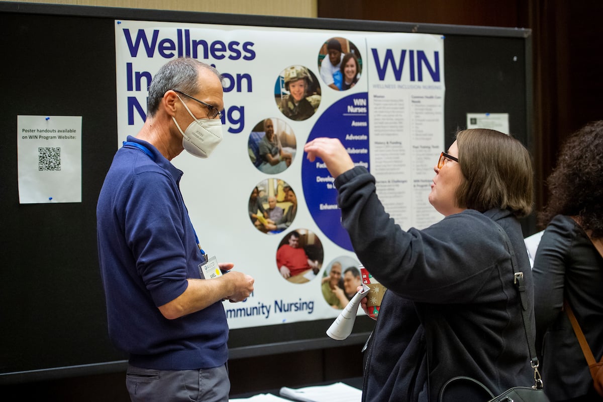 Two people stand at an information table in front of a large display board titled “Wellness Innovation in Nursing” with a “WIN” header. One person wears a face mask and a name badge, while the other holds a sheet of paper and gestures while speaking; the board behind them includes photos, bullet points, and the words “Community Nursing,” suggesting a poster session or informational event.