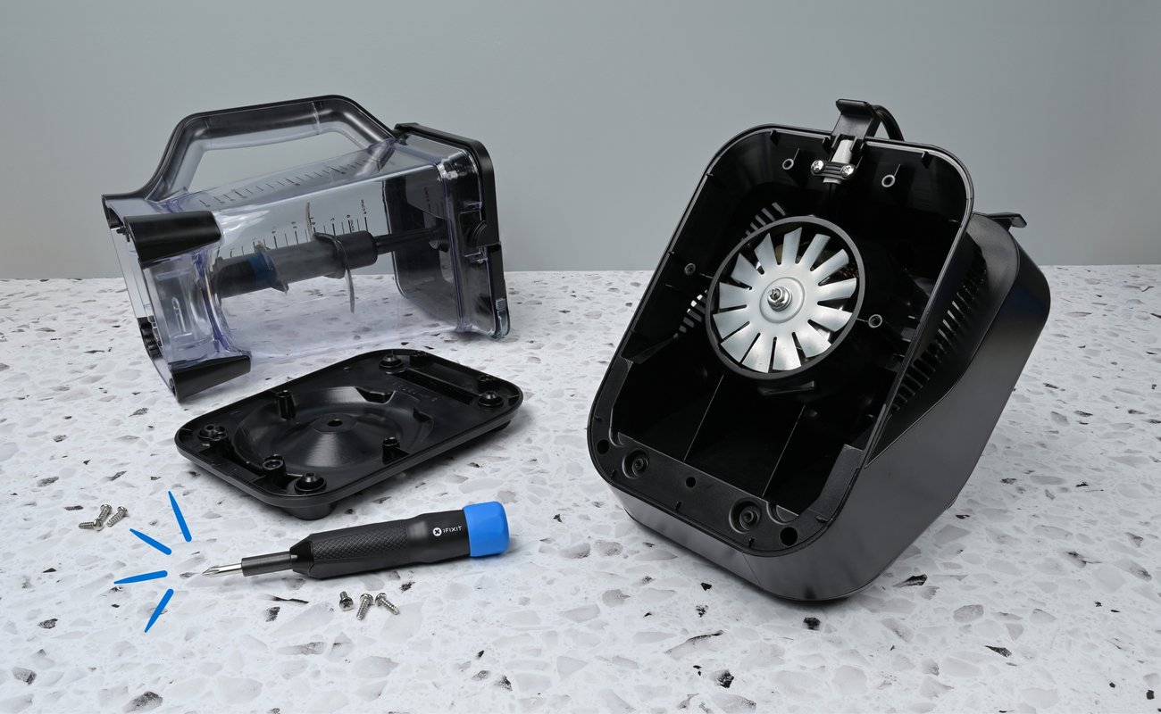 A disassembled black blender sits on a speckled countertop. The motor housing with a visible fan, the transparent pitcher, and the detached base plate are laid out, with the iFixit Narwhal Driver placed in front alongside several loose screws.