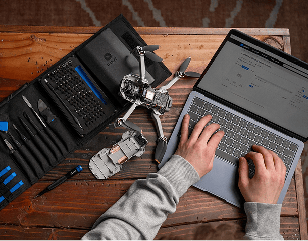 Top-down view of a person working on a laptop next to a disassembled drone and an open iFixit Pro Tech Toolkit on a wooden table.