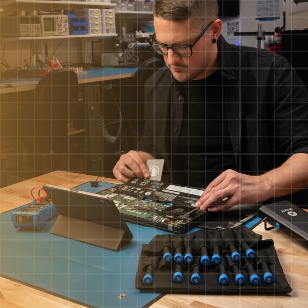 A technician works on an open laptop motherboard at a workbench, using iFixit tools laid out in front of him, with a tablet displaying repair instructions nearby in a professional workshop setting.
