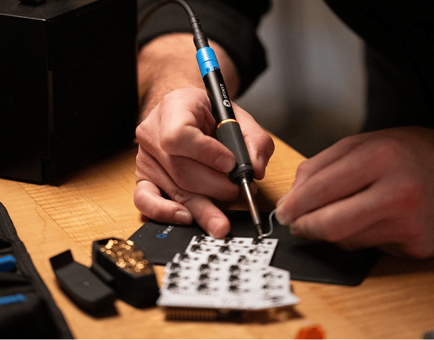 Close-up of hands using the iFixit Smart Soldering Iron to work on an electronic circuit board, with precision tools and soldering accessories on a wooden table.