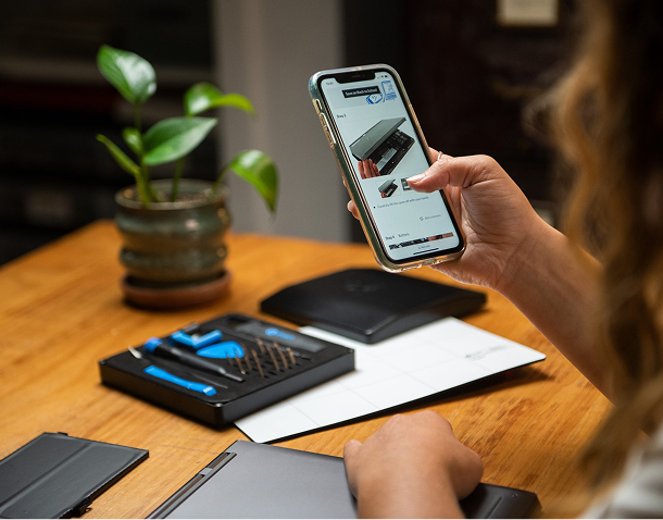 Close-up of a person using a smartphone to follow an iFixit repair guide while working on an electronic device, with an Essential Electronic Toolkit and Magnetic Project Mat on a wooden table.