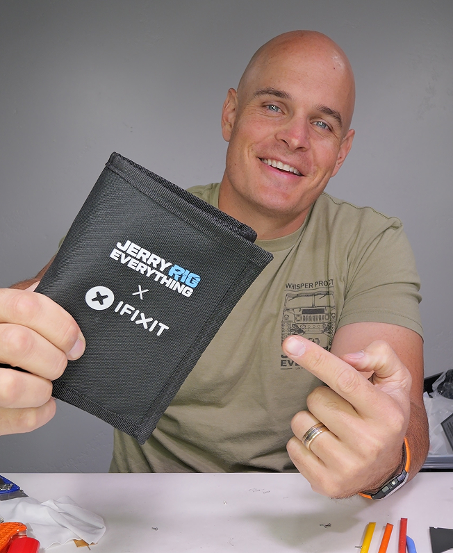 A smiling man at a workbench holds up a black JerryRigEverything × iFixit toolkit case toward the camera, with small repair tools scattered on the white table in front.
