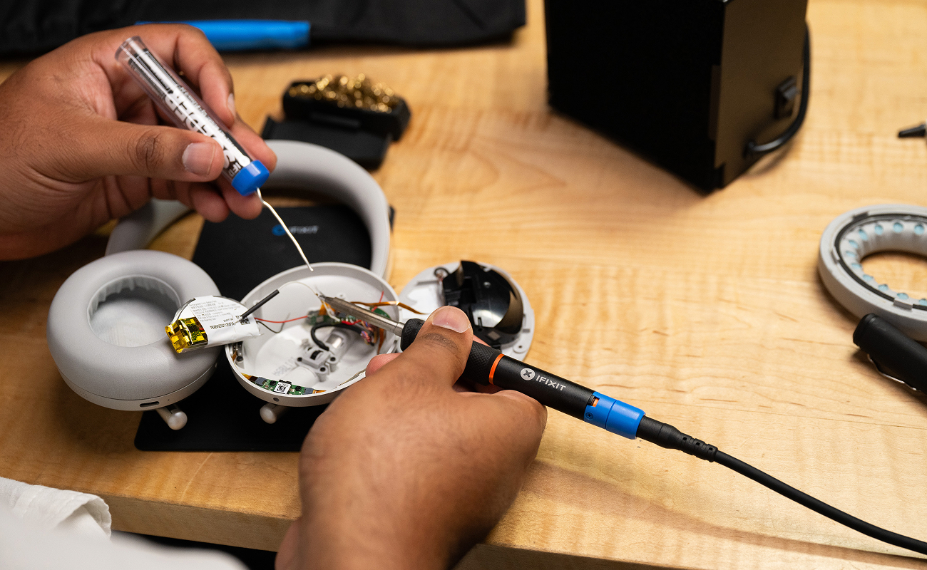 A close-up of hands repairing a pair of over-ear headphones on a wooden workbench using the iFixit FixHub Portable Soldering Station. One hand holds the soldering iron while the other feeds solder wire, with internal components and a battery visible inside the opened ear cup.