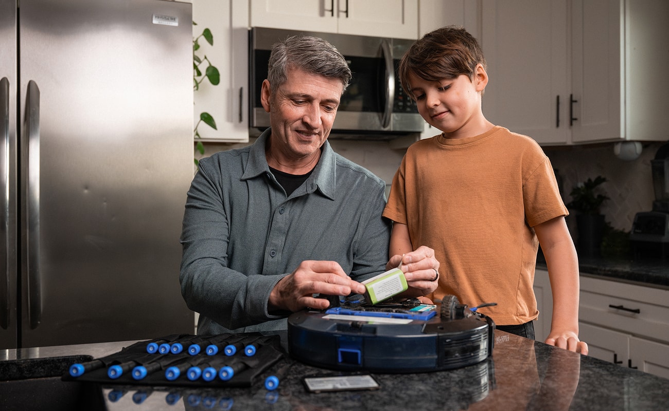 An adult and a child stand at a kitchen counter while working on an opened robot vacuum, with repair tools laid out in the foreground.