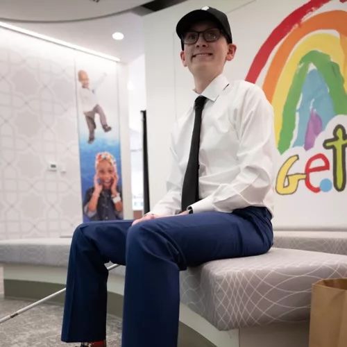 Image of a young man sitting on chair, wearing a black ball cap and a white shirt with a tie. 