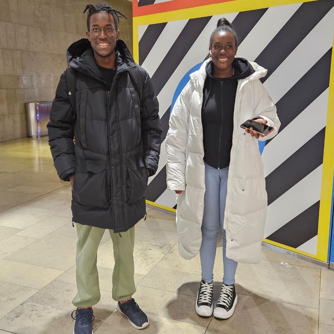 Image of a man and woman wearing coats in front of a black and white sign. 