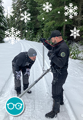 Two people standing on a snowy road holding a metal bar and measuring snow depth. Illustrated snowflakes appear on the top and an eyeglass graphic appears on the bottom left.