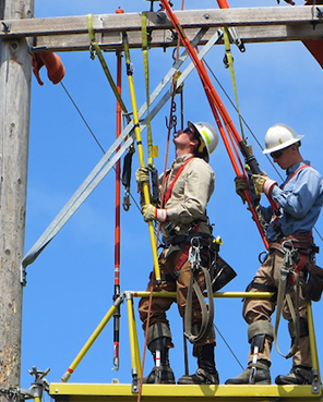 Two utility lineworkers in safety gear working on electrical equipment while suspended on a platform attached to a power pole. 