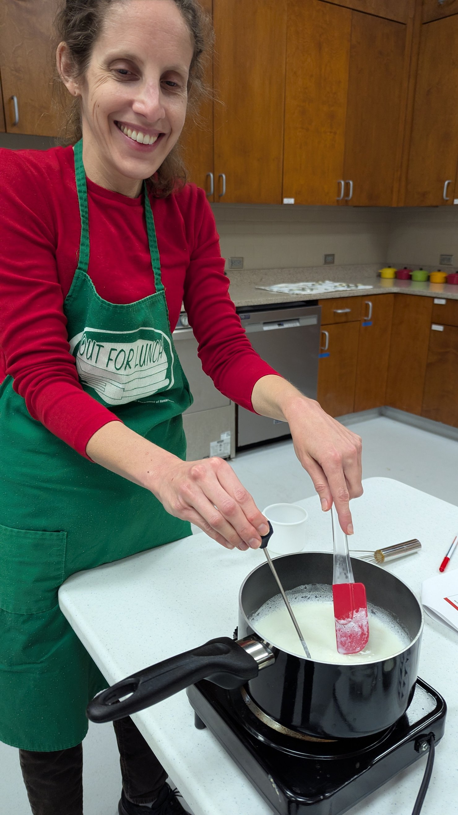 A woman in a red shirt and green apron checks the temperature of her yogurt using a food thermometer.