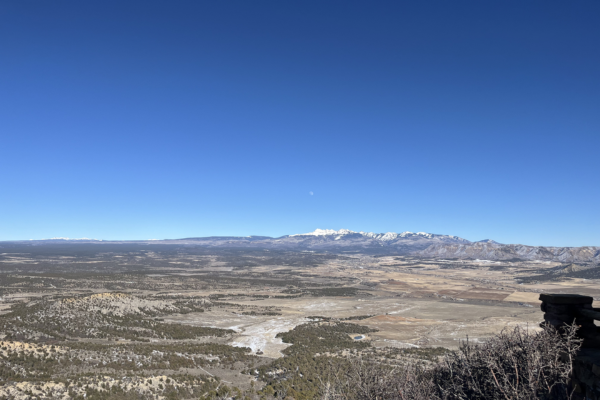 Landscape photo of a mountain range with some snow on top In the foreground theres desert and fields with some trees