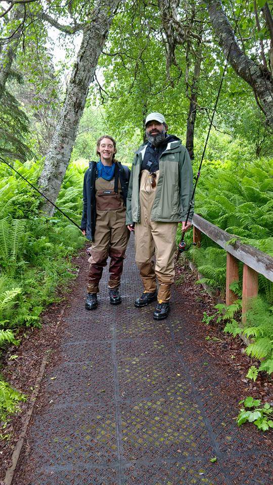 Abby and Andrew in waders holding fishing rods standing on a forest path surrounded by lush green ferns and trees in Alaska.