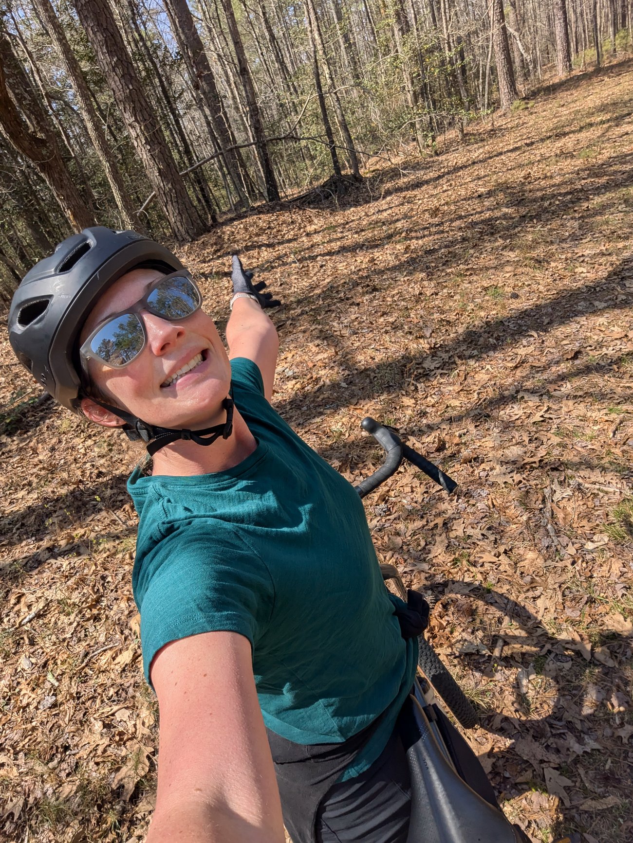 Selfie of Abby wearing a bike helmet and sunglasses on a wooded trail, pointing into the forest while standing with her bike