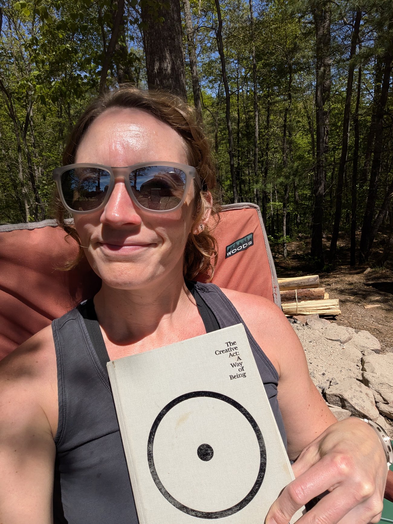 Abby sitting outdoors in a camp chair wearing sunglasses, holding the book *The Creative Act: A Way of Being*, surrounded by trees and sunlight