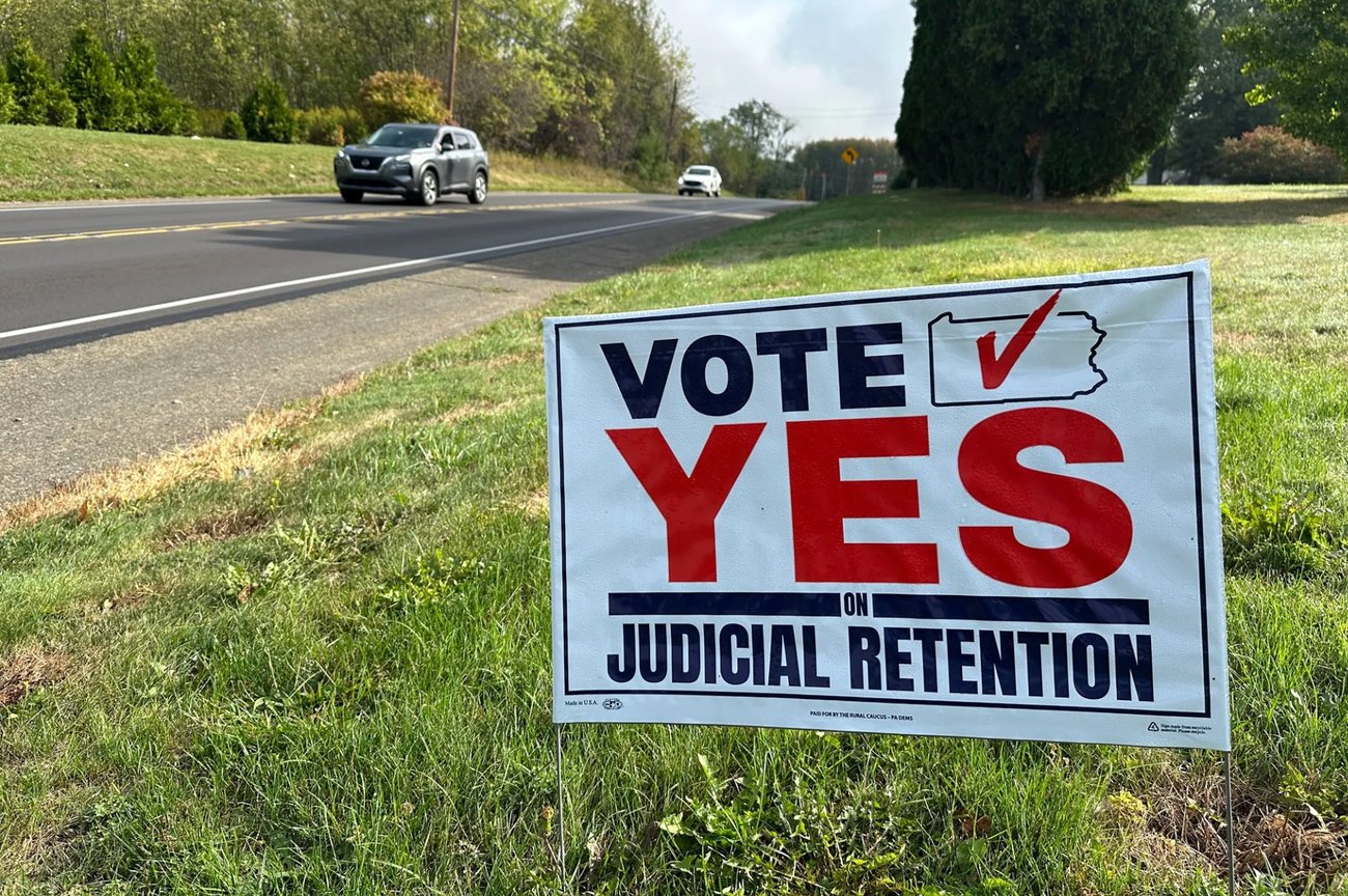 Yard sign along road, reading "Vote Yes on Judicial Retention" with an illustration of the outline of Pennsylvania with a big red checkmark atop it