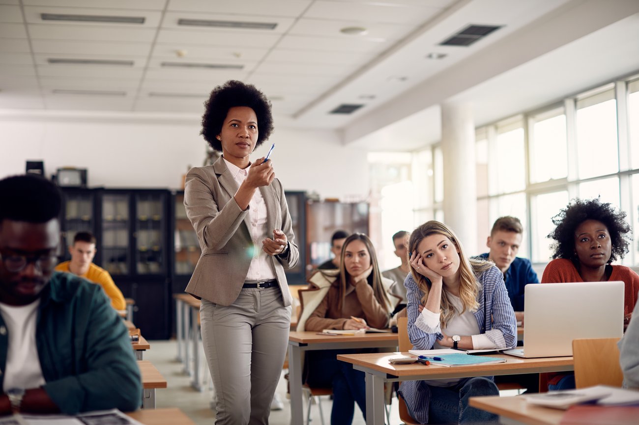 An lecturer stands among students seated in a classroom, drawing student attention towards the front of the room. An lecturer stands among students seated in a classroom, drawing student attention towards the front of the room.