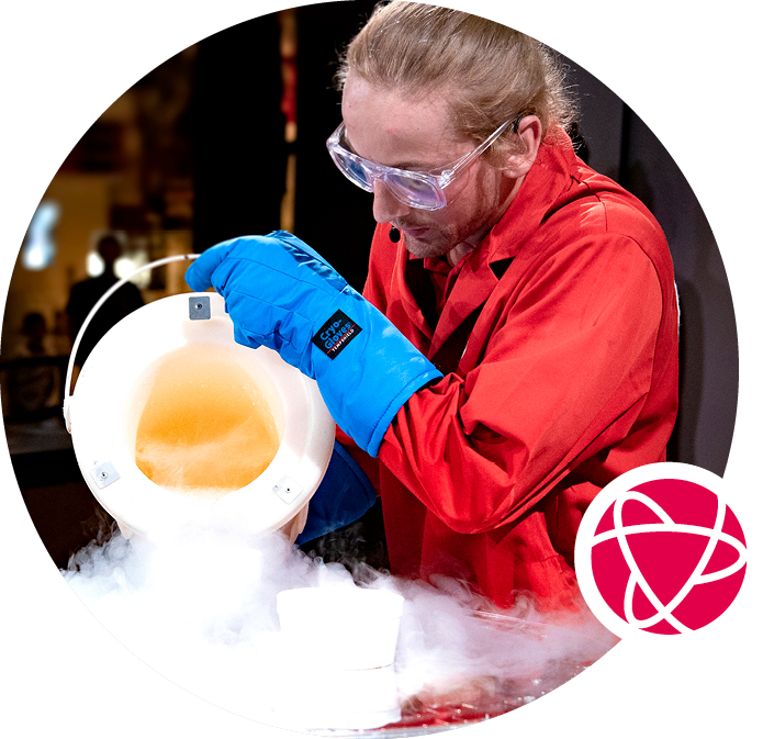 A museum guide in a red labcoat pours a liquid from a white bucket, creating a white fog.