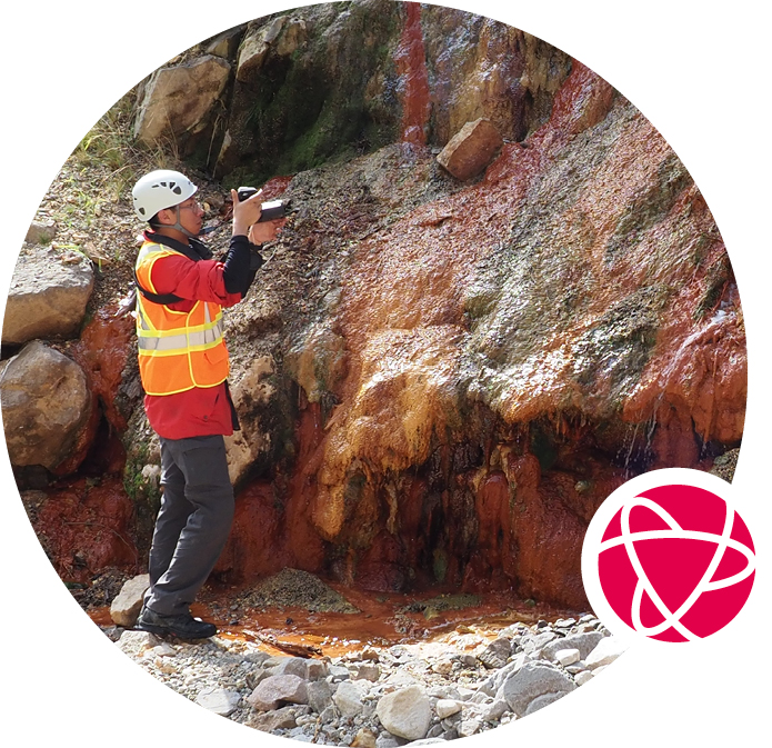 A worker in an orange safety vest and helmet points a survey instrument at a wall of red and brown rocks.