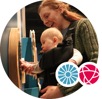 A woman holds her baby in a carrier on her front. She holds the baby's hand, helping them touch a textured display on the wall.