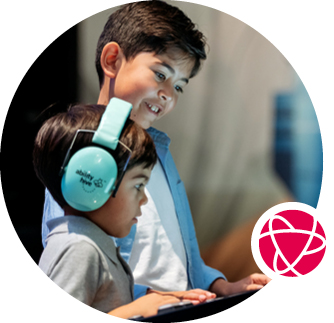 Two children smile while looking at a museum interactive. The younger child wears noise-reduction headphones.