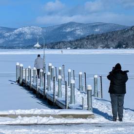person taking a picture of another person on dock on lake george in winter