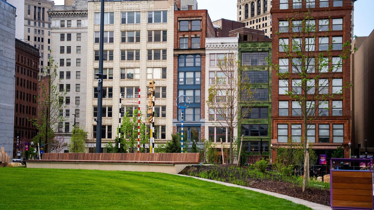 A city park with a large green lawn and landscaped plants in the foreground, surrounded by tall office buildings and apartments in the background.