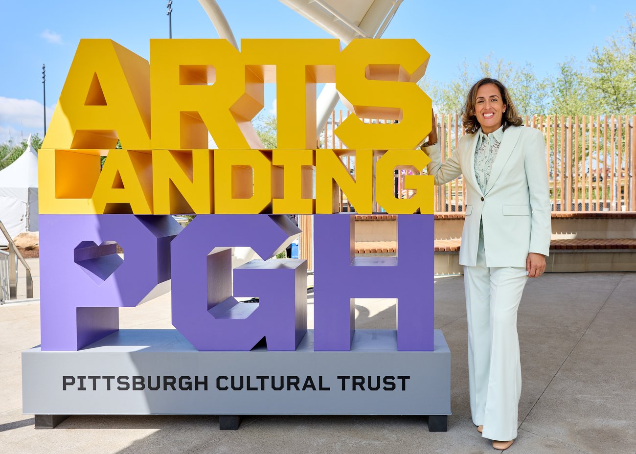 A woman in a light-colored suit smiles and stands next to a large, colorful ARTS LANDING PGH sign. The base reads PITTSBURGH CULTURAL TRUST. There are trees and a fence in the background.