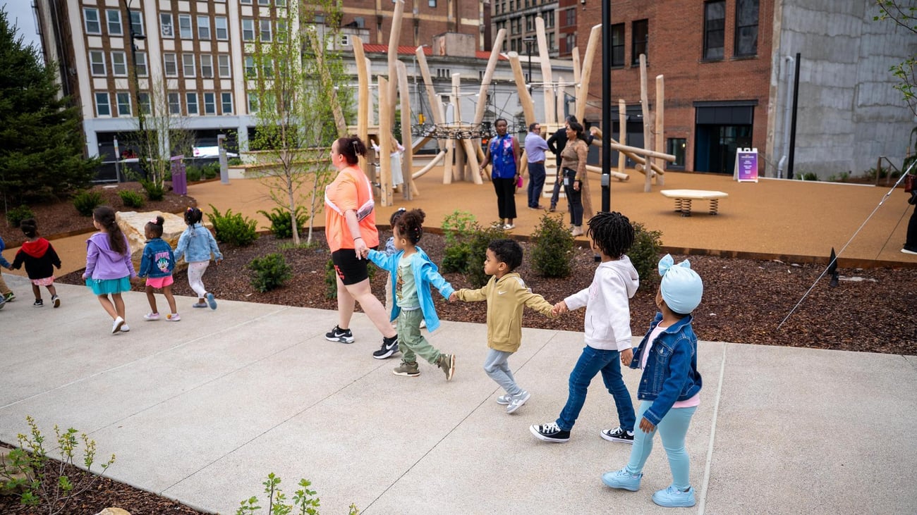 A group of young children holding hands walk in a line outdoors, led by an adult, near a modern playground with climbing structures and several adults in the background. The scene is set in an urban park. A group of young children holding hands walk in a line outdoors, led by an adult, near a modern playground with climbing structures and several adults in the background. The scene is set in an urban park.