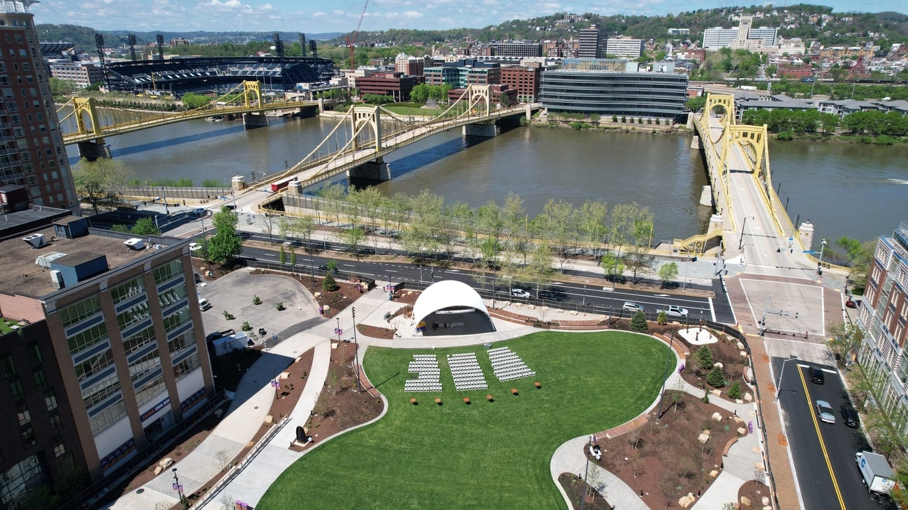 Aerial view of Arts Landing with a small amphitheater, two yellow bridges crossing the river, and city buildings and hills in the background. Aerial view of Arts Landing with a small amphitheater, two yellow bridges crossing the river, and city buildings and hills in the background.