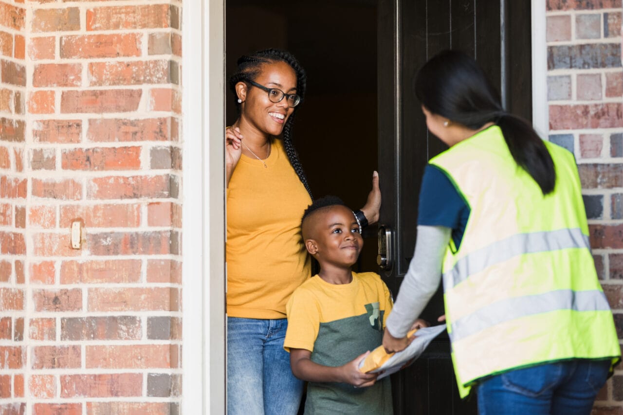 A boy receives a package from a delivery person at the front door in front of his mom. 