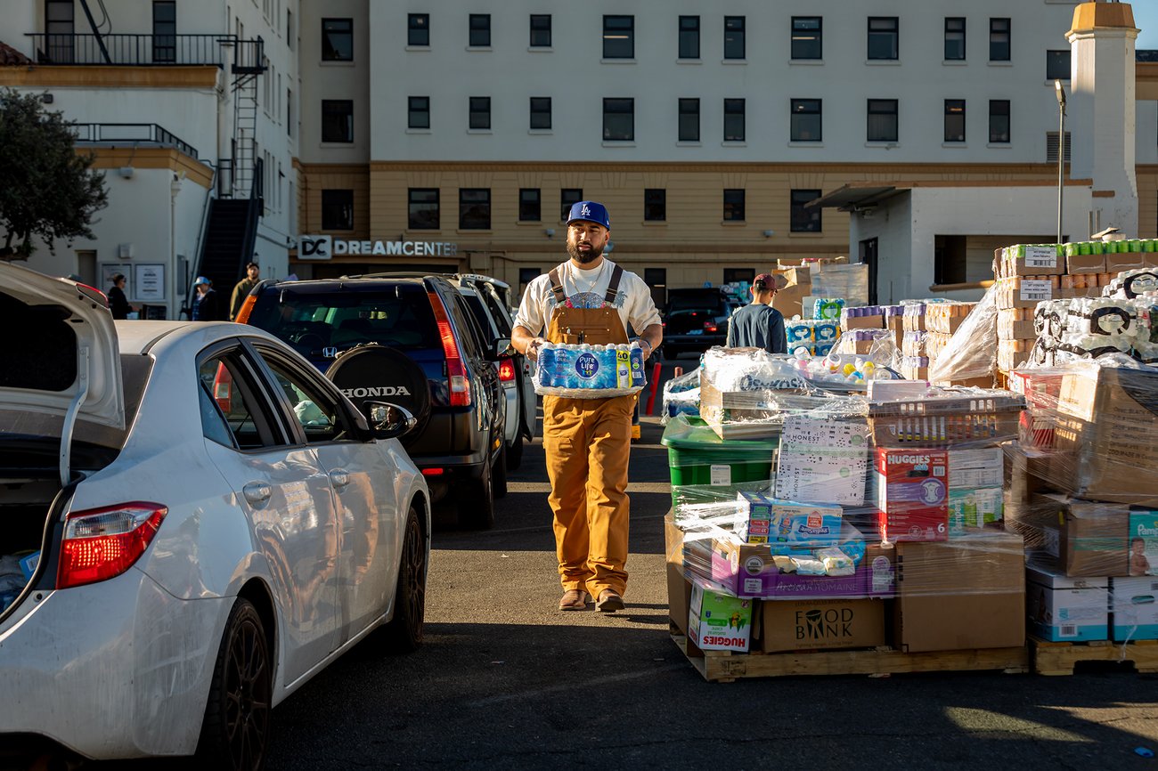 A person wearing light brown overalls and a blue Los Angeles baseball cap carries a crate of bottled water and walks towards a white car with the trunk open as it waits in line to receive donations at a donation site. A person wearing light brown overalls and a blue Los Angeles baseball cap carries a crate of bottled water and walks towards a white car with the trunk open as it waits in line to receive donations at a donation site.