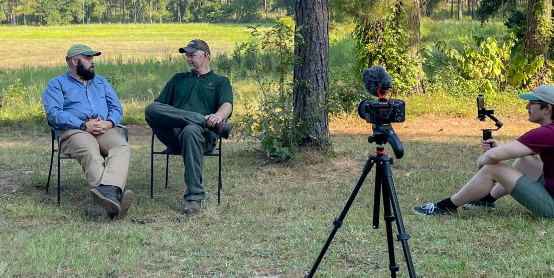 A wide shot of an outdoor interview taking place in a grassy field bordered by trees. Two men are seated in chairs on the left, engaged in conversation; one wears a blue button-down shirt and a tan cap, while the other is in a dark green polo and a brown cap. In the foreground on the right, a camera with a large fuzzy windscreen (deadcat) is
mounted on a tripod, pointing toward the men. A third person sits on the ground nearby, operating a handheld gimbal with a smartphone to capture additional footage.