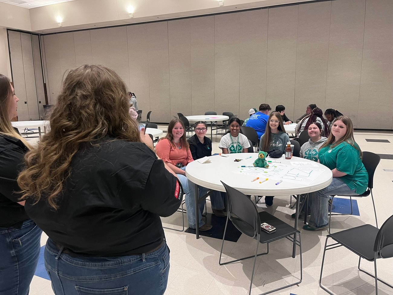 Teen State 4-H officers snap a photo of South Central district teen 4-H'ers posing at a table with Clover, a small, green plush bear as the centerpiece