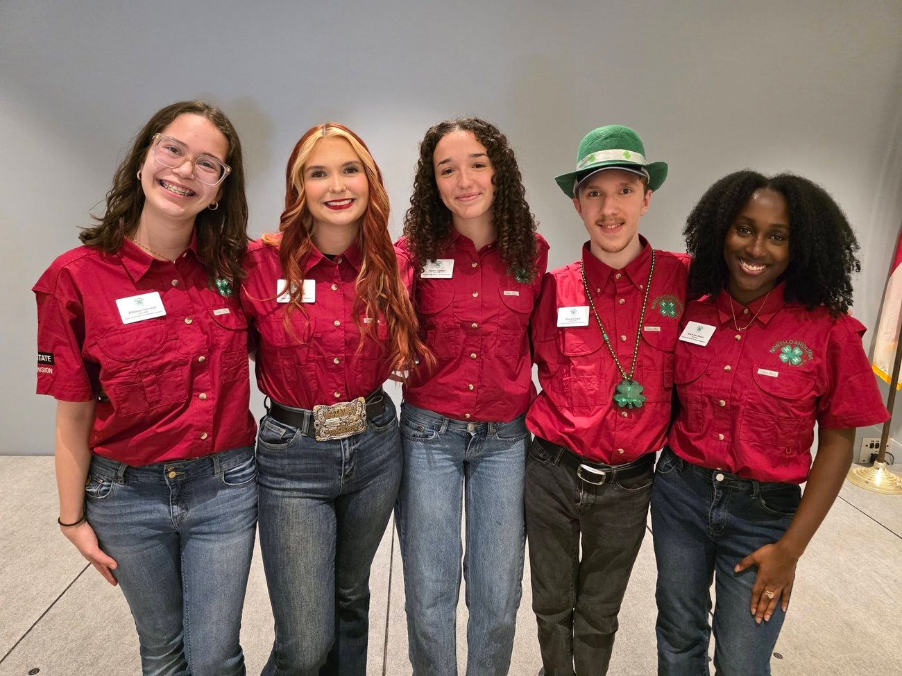 4 teen 4-H'ers, posed in a line with their arms around one another, dressed in matching red shirts with 4-H clover logos