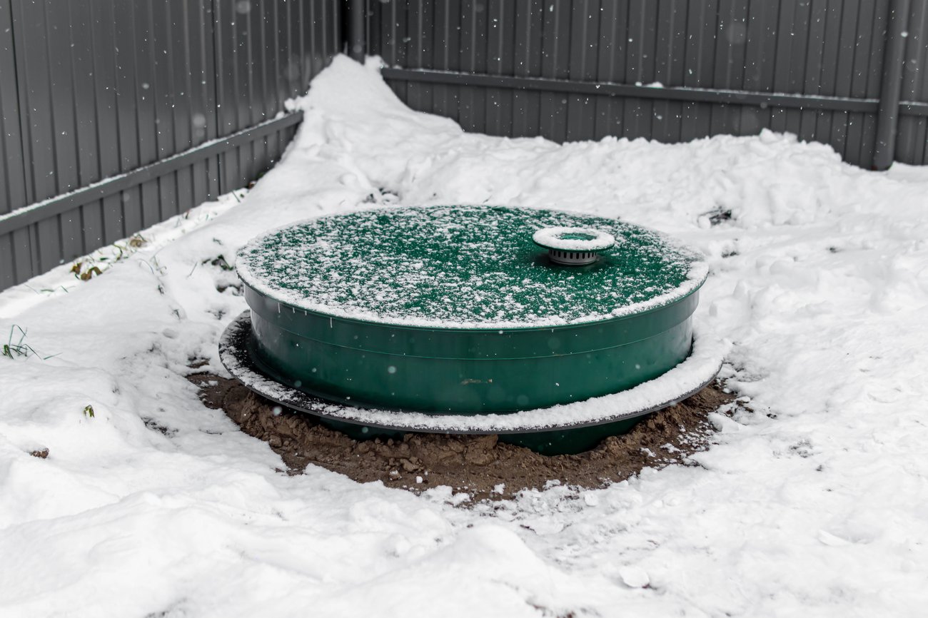 The exposed access port of a septic tank in a yard covered in snow.