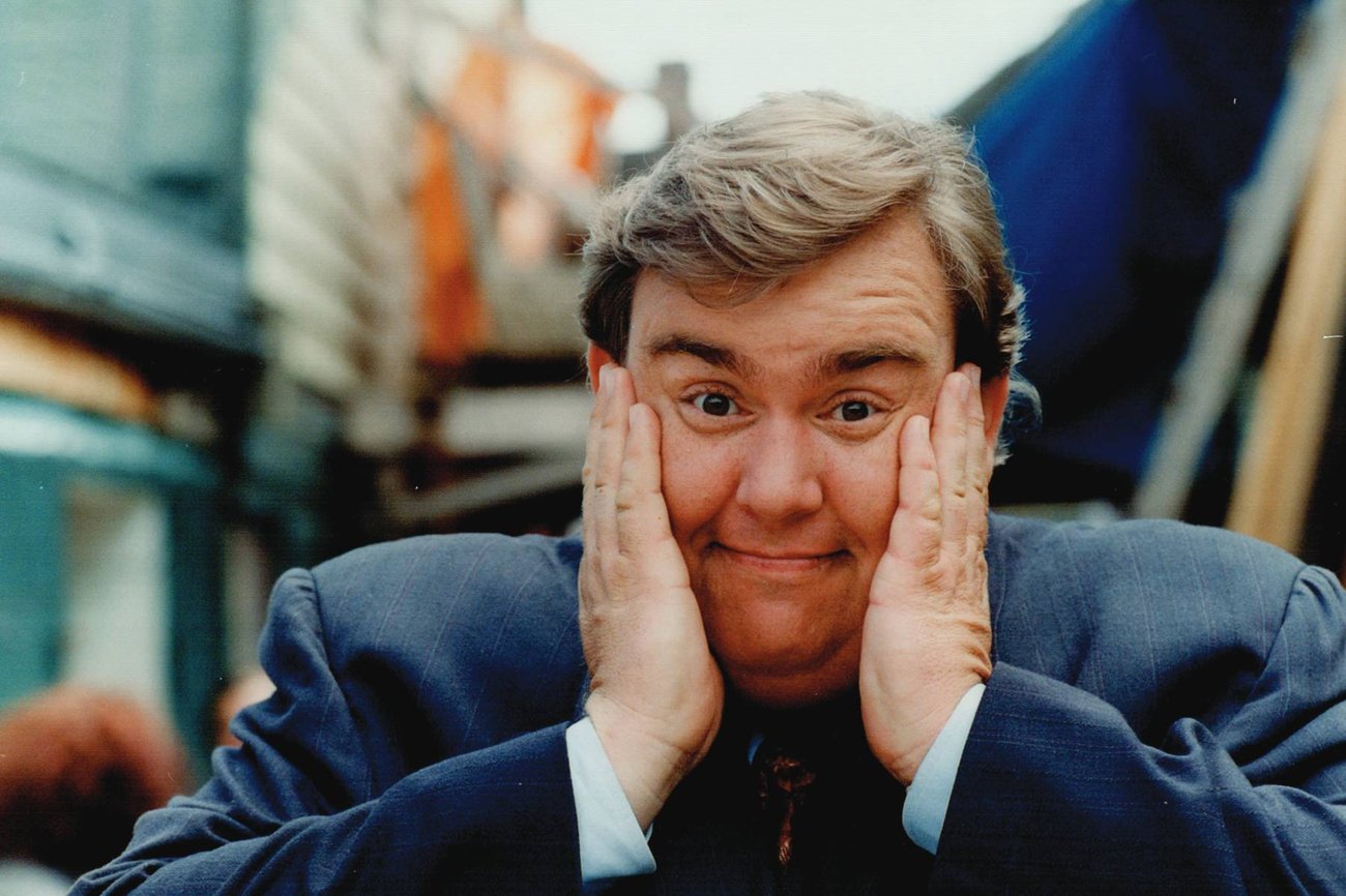 Image of actor John Candy smiling with his hands pressed against his cheeks.