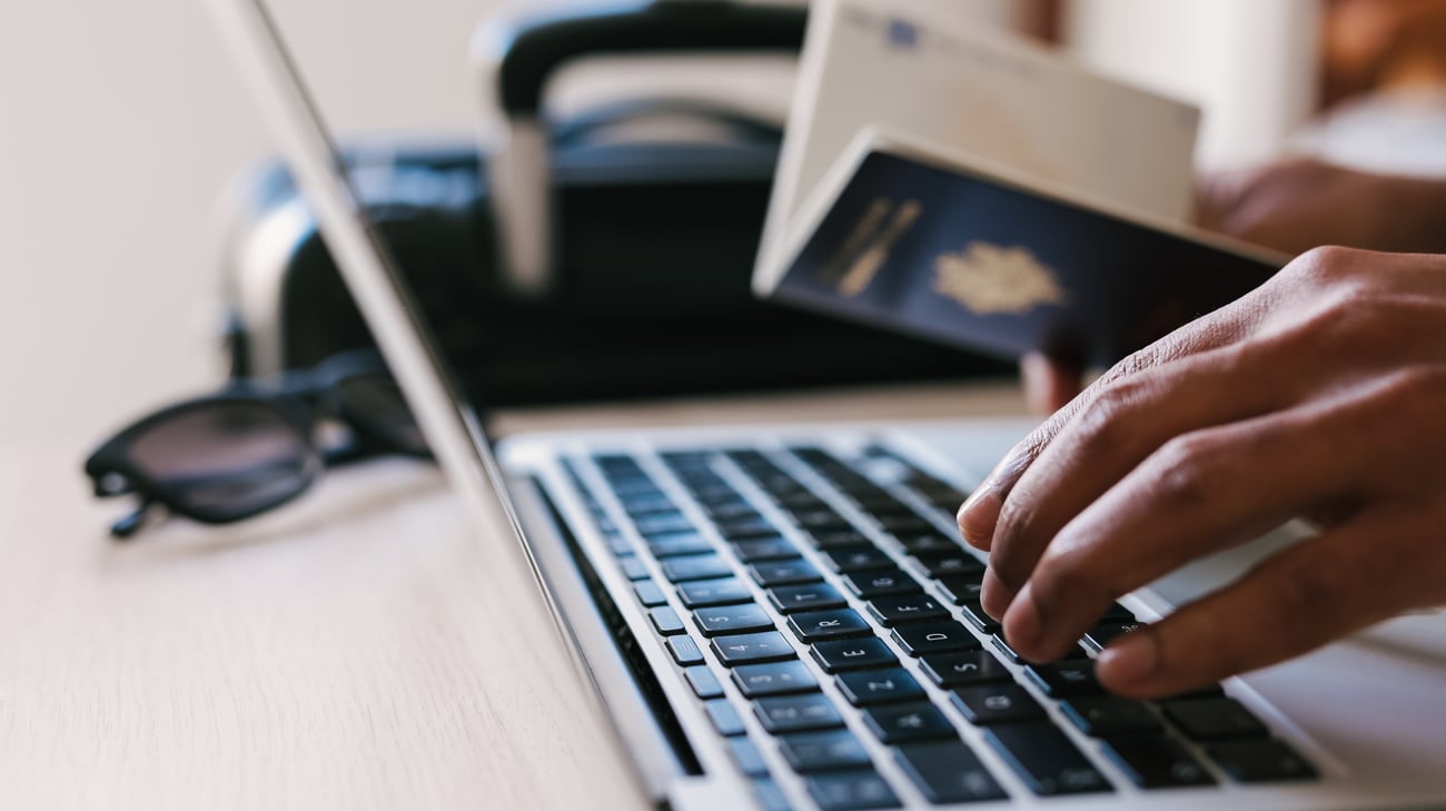 Person holding passport typing on computer. In the blurred background, there is a piece of luggage and sunglasses