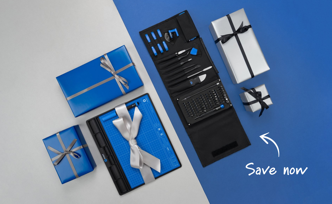 A flat lay of iFixit repair gifts on a split gray and blue background, featuring the FixMat wrapped with a silver ribbon and the open Pro Tech Toolkit roll displaying neatly organized tools and bits. Blue and silver gift boxes surround the products, with a handwritten arrow and text reading “Save now” adding a festive promotional touch.