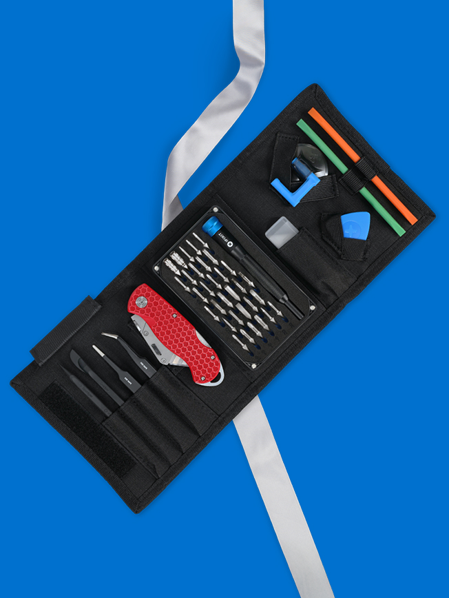 The iFixit X JerryRigEverything Toolkit opened on a bright blue background, showing an array of precision tools, bits, tweezers, opening tools, and a red utility knife neatly arranged in the black roll. A silver ribbon runs through the composition, giving the professional toolkit a festive, gift-worthy touch.