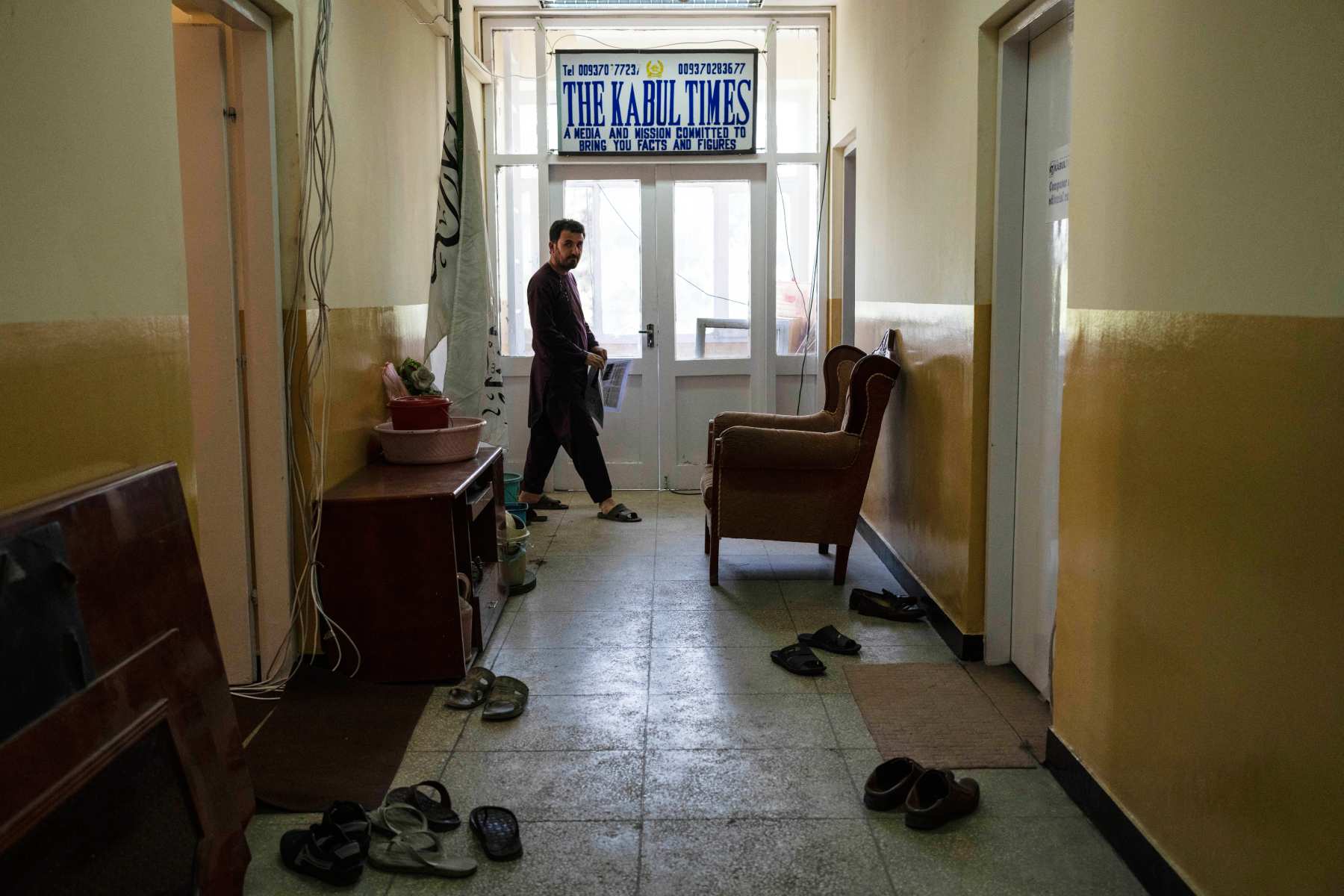 A hallway in a building with yellow walls and tiled floor. A man walks past a sign that read The Kabul Times.