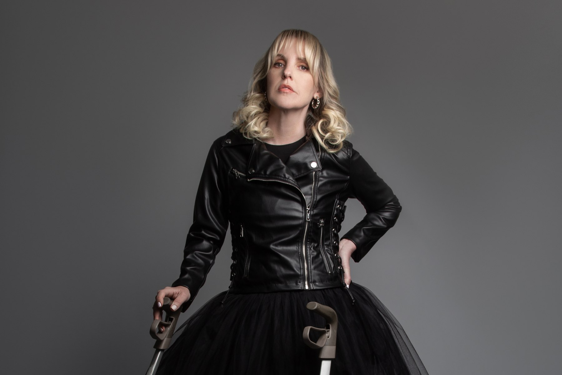 A photo of a blonde woman in a leather jacket and black tulle skirt holding the handles of a mobility device.