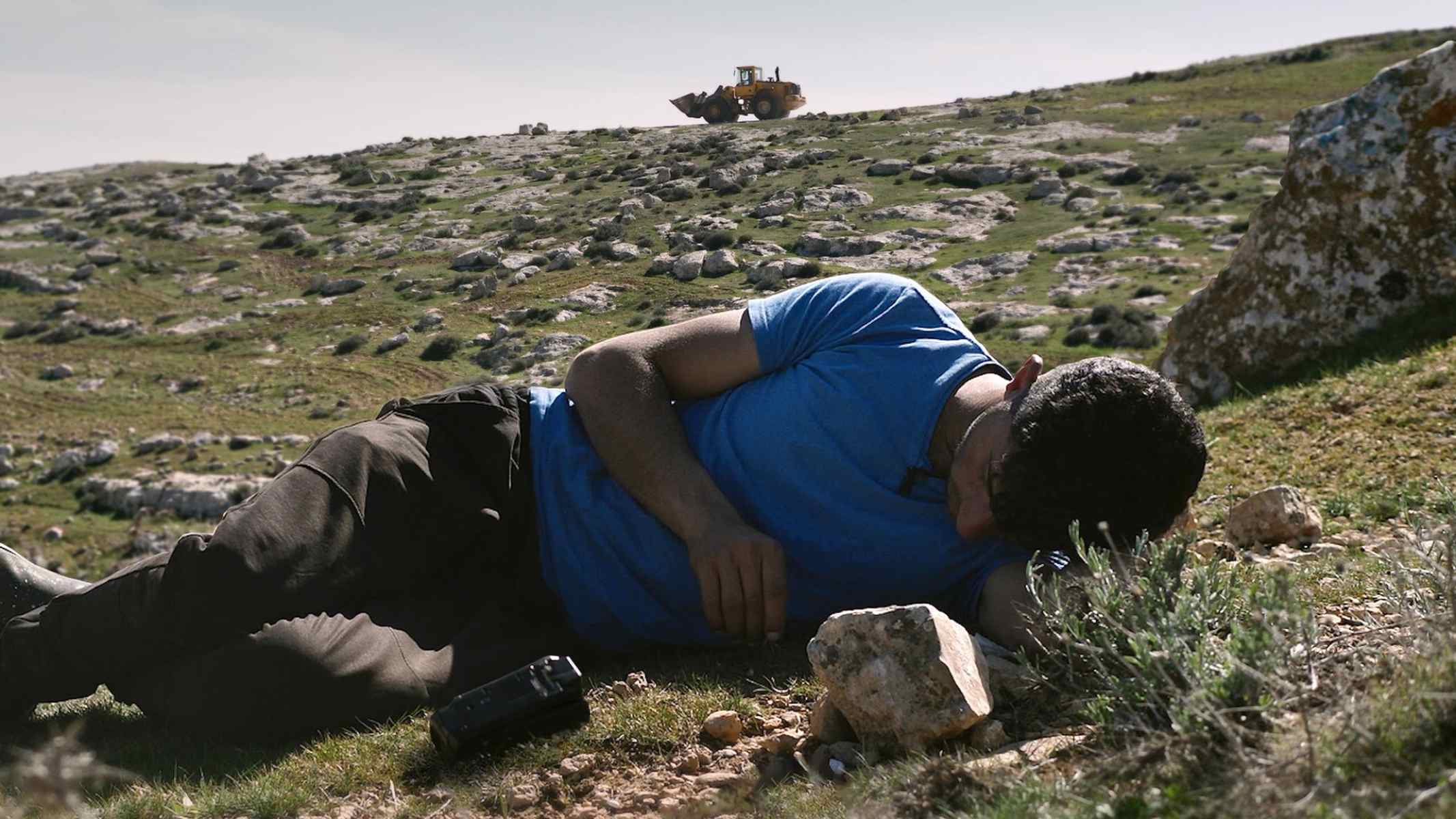 A man in blue shirt lies down on a hilly green field covered in large rocks. A construction vehicle can be seen on the horizon.