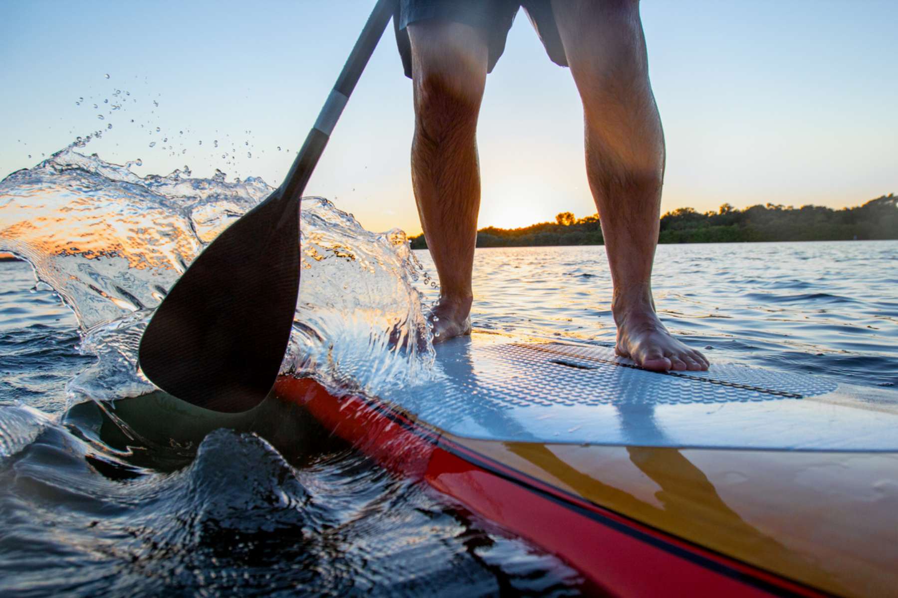 A close up of man on a paddleboard at sunset. He's pushing the paddle forward with water spraying forward to the camera.