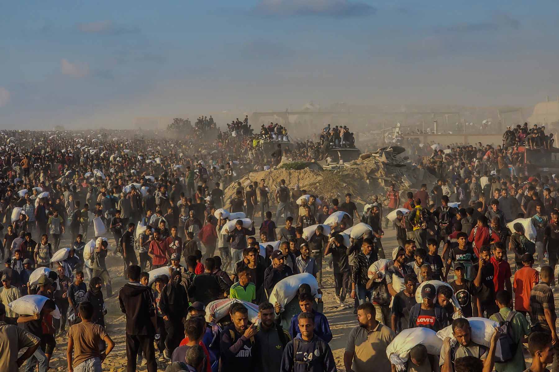 Under a hazy sky, thousands of people, some walking alone, others clustered in groups, some with vehicles, carry heavy white sacks on their shoulders or heads
