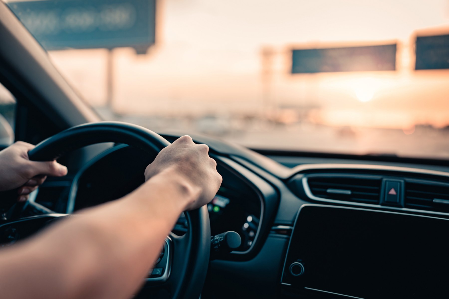A photo of a man's hands on steering wheel with a view of the highway ahead and setting sun through the window.