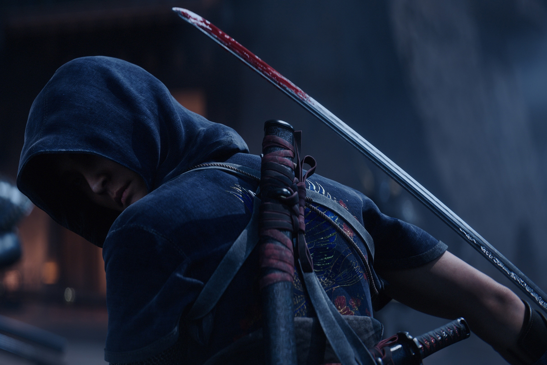 Image shows a hooded figure with their back to the camera turning around, holding a weapon in one hand and on their back.